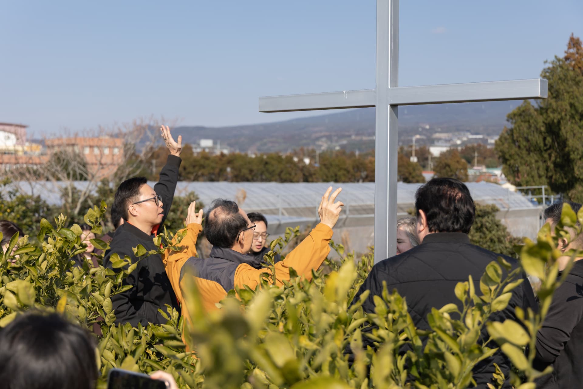 People gathered in prayer around a white cross on Jeju land, with mountains and sky in the distance.